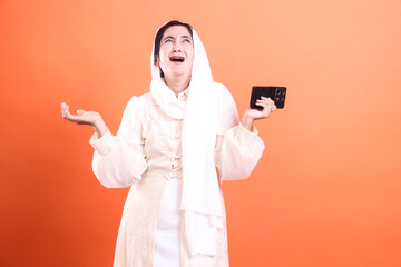 Frustrated Muslim Woman with Smartphone: A Tech Disappointment Portrayal on Orange, conveying frustration and technology issues, isolated on a orange background.