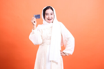 Modern Muslim Woman Holding Credit Card: A confident Muslim woman showcases her financial empowerment with a debit card, isolated on a orange background.