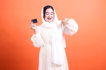 Joyful Muslim Woman Holding Credit Card: Capturing the ease and security of modern finance with tradition gracefully, isolated on a orange background.