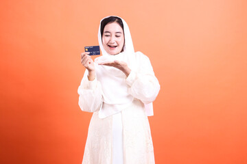 Happy Muslim Woman Presenting Credit Card: Embracing Modern Finance with a Smile on Orange, reflecting financial empowerment.