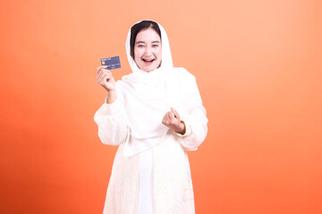Cheerful Muslim Woman with Credit Card: Empowered Shopping Experience. Isolated on an orange background, radiating positivity and financial freedom.