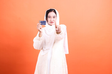 Muslim woman presenting debit card with caution: shop smart and stay safe online, isolated on a orange background.