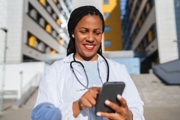 african american woman doctor on break use cellphone in front hospital