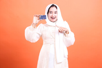 Cheerful Muslim Woman Presenting a Credit Card with Excitement, symbolizing financial opportunity, isolated on a orange background.