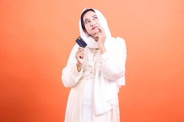 Pensive Muslim Woman with Credit Card: Planning Her Next Purchase. Isolated on a orange background,...