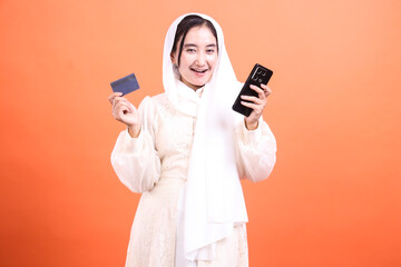 Happy Muslim Woman Shopping Online with Smartphone and Credit Card, showcasing modern Islamic finance and technology, isolated on a orange background.