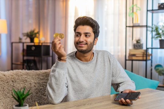 Young Indian man with bitcoin checks cryptocurrency exchange charts on smartphone app calculates approximate profits, looking on BTC coins in hand celebrating win. Hispanic guy looks satisfied at home