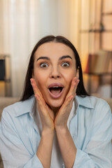 Young woman at home on sofa raising eyebrows, opening mouth with amazed surprised look wow seeing...