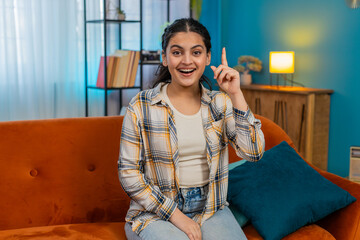 Indian young woman sitting on sofa raising index finger, sudden Eureka expression, joyful...