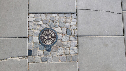 Circular metal drain cover set into a cobblestone pattern surrounded by gray concrete slabs,...