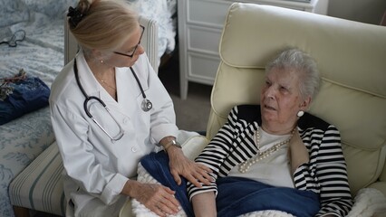 A mature woman sits comfortably in a chair at home while a healthcare professional speaks with her, offering companionship and medical advice during the visit.