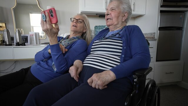 An elderly woman in a wheelchair sits with her daughter, sharing memories and joy through a smartphone while enjoying a warm visit at home.
