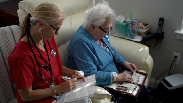 A caregiver in scrubs records medication details while an elderly woman looks through photo albums in a comfortable living room, showcasing companionship and care.