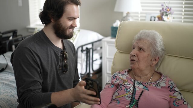 A young man smiles as he shows his grandmother pictures on his smartphone. They are sitting together in a warm living room filled with love and memories.