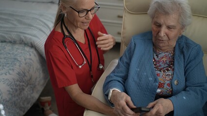 A healthcare worker helps an elderly woman with a remote control while seated in a cozy room,...