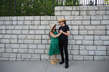 woman in green dress in hat and man in hat are near brick wall,