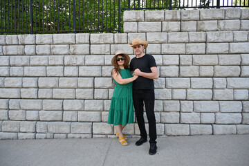 woman in green dress in hat and man in hat are near brick wall,