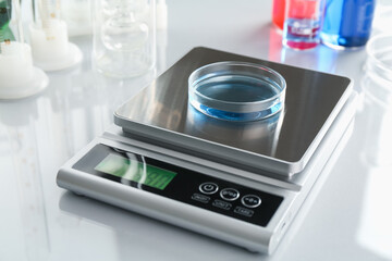 Scale with Petri dish of sample and other laboratory glassware on white mirror table, closeup
