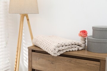 Woolen blanket, boxes, beautiful flower in vase on wooden chest of drawers and lamp near white wall...