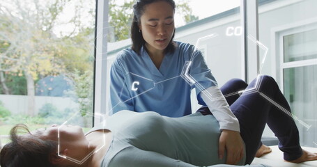 Physiotherapist in scrubs raising patient's bent leg on table in clinic by windows with AR overlays