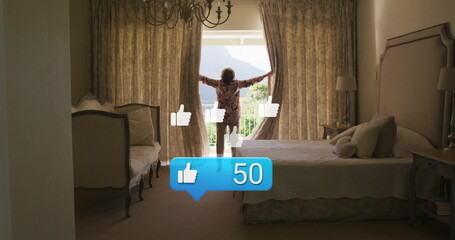 Pulling back floral curtains senior woman in floral robe at window in bedroom, showing balcony view