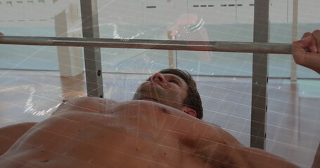 Lifting shirtless mid adult male bench pressing in gym, with barbell and floor-to-ceiling windows