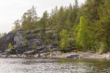 Two kayaks lie on the shoreline of a rocky forest peninsula, with pine trees and calm water under...