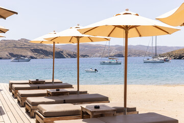 Lounge chairs on the beach with umbrellas ready for guests on small Greek island with views of blue sea and boats. 