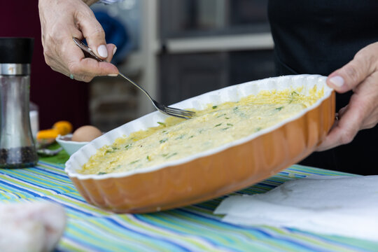 Woman smoothing a Greek potato pie in a ceramic dish with festive blue and green striped tablecloth.