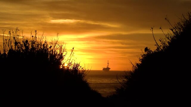 An offshore oil rig is silhouetted against an orange sunset and framed by waves and shoreline vegetation by the UCSB campus near Santa Barbara, California.