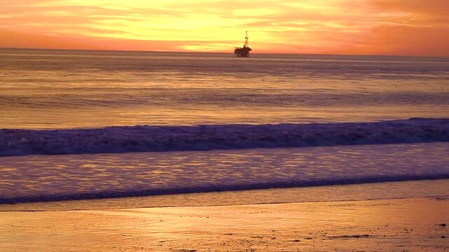 Purple and orange waves pound the beach where an offshore oil drilling platform is silhouetted against the sunset by the UCSB campus near Santa Barbara, California.