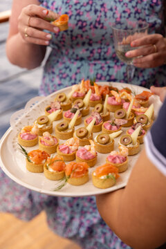 Middle aged woman in floral dress on a cruise holding a glass of white wine getting offered a plate of seafood and cheese hor d'oeuvres by a staff member. 