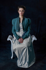 A Regency woman wearing a blue cotton dress and short spencer with soutache braid sitting against a...