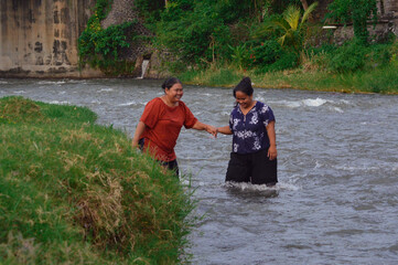 A woman gently holds her friend's hand, helping her find courage to step into the cool river as...