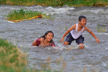 Fototapeta premium A young boy shows a joyful expression while playing with a girl in the flowing river, surrounded by lush grass and wild riverside plants.