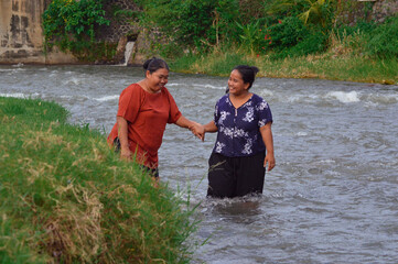A smiling woman holds her friend's hand, inviting her into the refreshing stream as they both laugh...