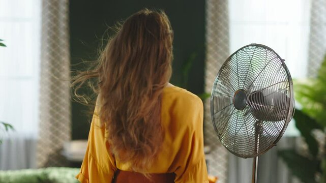 Viewed from a rear angle, a woman with long wavy brown hair stands before a spinning electric fan in a bright, airy living space, finding comfort and relief from summer heat.