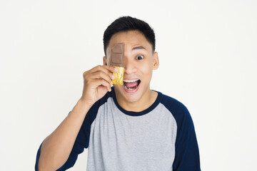 Young man holding golden-wrapped chocolate bar over his eye, expressing playful enthusiasm. Ideal image for themes of happiness, fun, sweets, and youthful enjoyment.