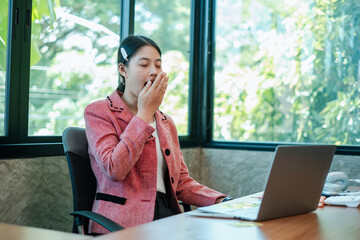 A young professional woman yawning at her desk in a modern office setting, surrounded by large...