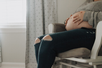 Expectant mother sitting in a white chair at home, wearing gray sweater and ripped blue jeans,...