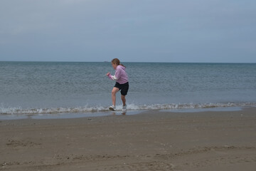 Young girl playfully runs along the shoreline, splashing in the shallow waves. The ocean stretches into the distance under a cloudy sky, capturing a carefree beach moment.