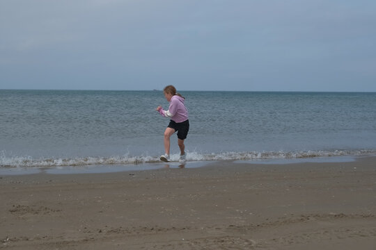 Young girl playfully runs along the shoreline, splashing in the shallow waves. The ocean stretches into the distance under a cloudy sky, capturing a carefree beach moment.