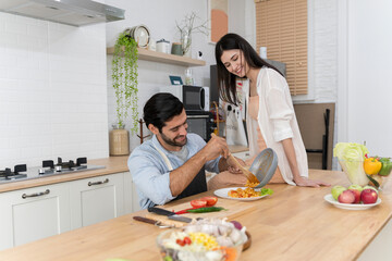 Man serving pasta onto plate while woman smiles beside him, happy couple preparing meal in modern kitchen