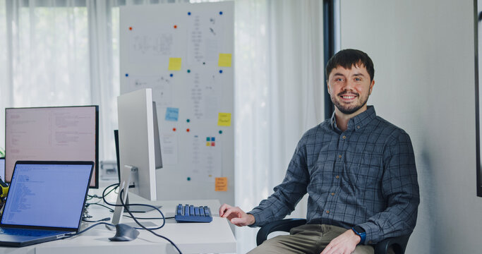 Portrait confident smiling male programmer working with robotics code AI software and looking at camera in tech office. Artificial intelligence dashboard, robotics, user interface, machine learning.