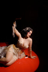 Young Caucasian woman in ornate golden corset, lying on red round stage floor, under hanging beads, dark background, front view. Concepts: cabaret, circus, retro, burlesque.