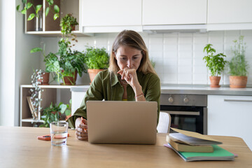 Pensive nervous woman working on laptop with books and water glass at home. Adult student managing deadlines, work pressures, remote job, prolonged screen time, stress businesswoman multitasking