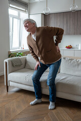 Senior man grimacing in pain attempting to stand up from couch due to spinal condition. Chronic osteochondrosis, vertebral degeneration, limited mobility, age related disorders, movement difficulties