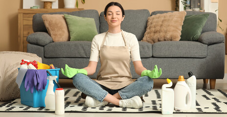 Young woman meditating on floor among cleaning supplies in living room