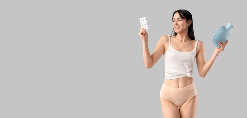 Young woman in period panties holding hot water bottle and pills on grey background with space for...