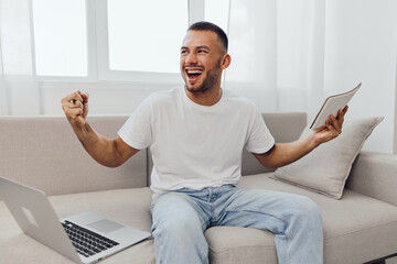 Happy man celebrating success at home with a laptop and notebook, showcasing enthusiasm and...